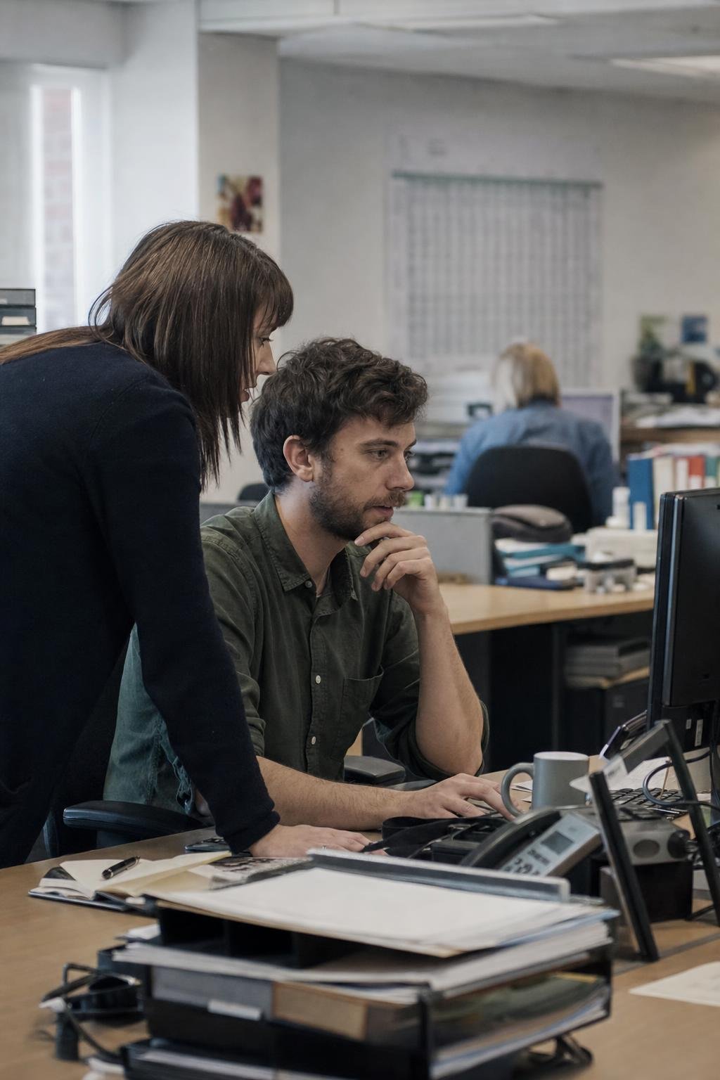 An engineer working at a borrowed desk inside a client's operations office, a colleague from the business leaning in to look at the screen with them.